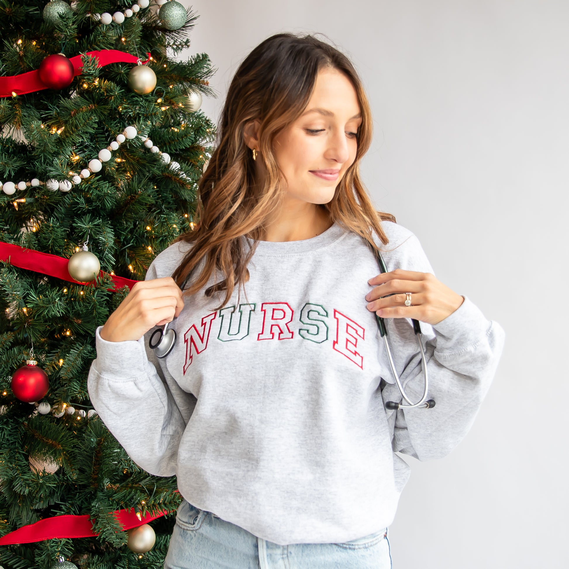 Woman styling an ash crewneck sweatshirt  that has the word NURSE embroidered in alternating red and green threads.