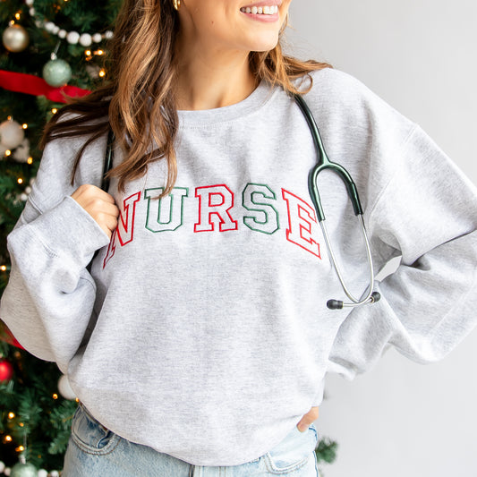 Woman wearing an ash crewneck sweatshirt that features curved text in red and green threads that says Nurse embroidered across the chest.