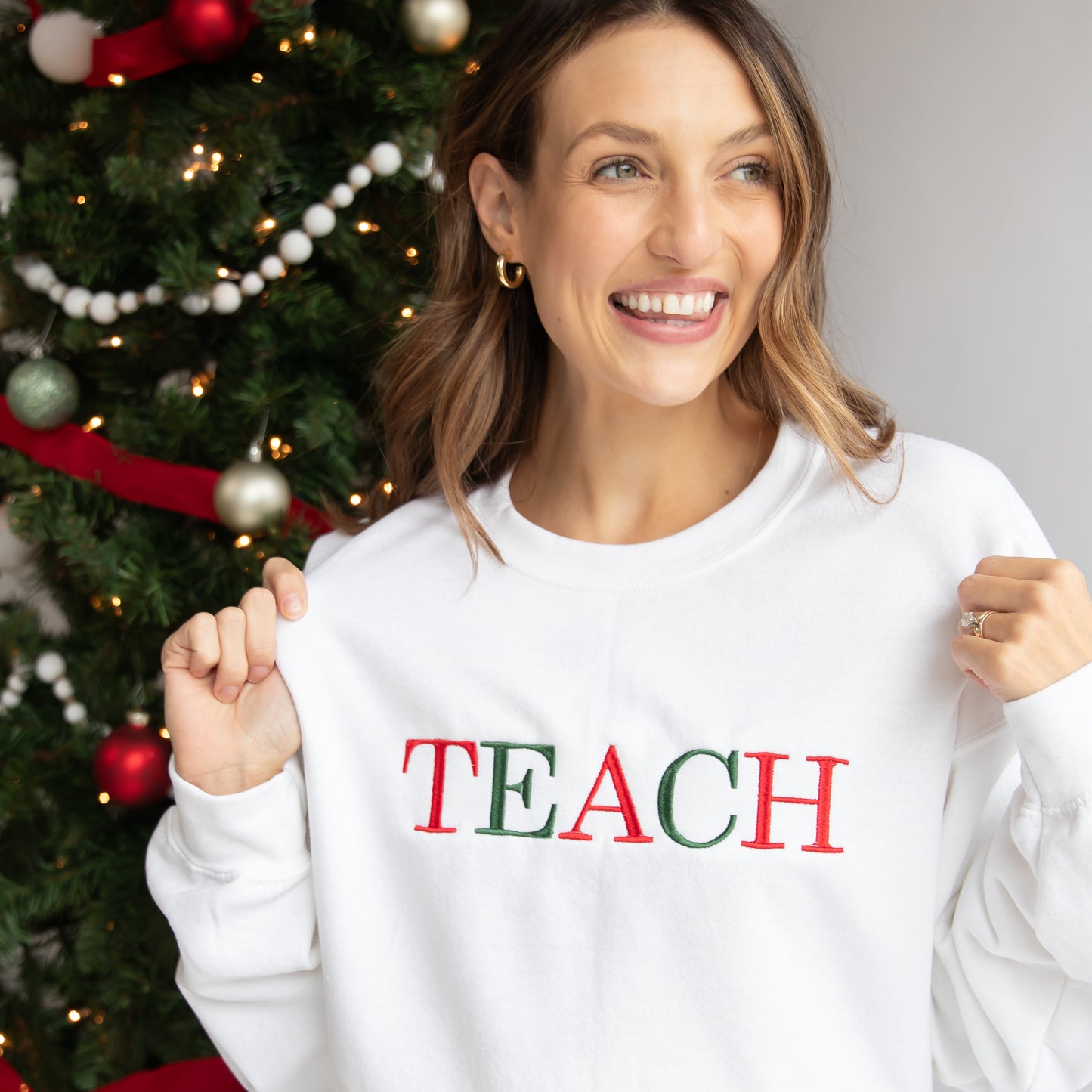 Woman standing in front of a Christmas tree wearing a white crewneck sweatshirt that has the word TEACH embroidered in alternating red and Green threads.