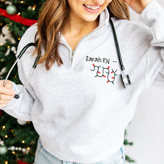 nurse standing by christmas tree wearing a light gray quarter zip with her name, credentials, and a christmas lights stethoscope design embroidered on the left chest