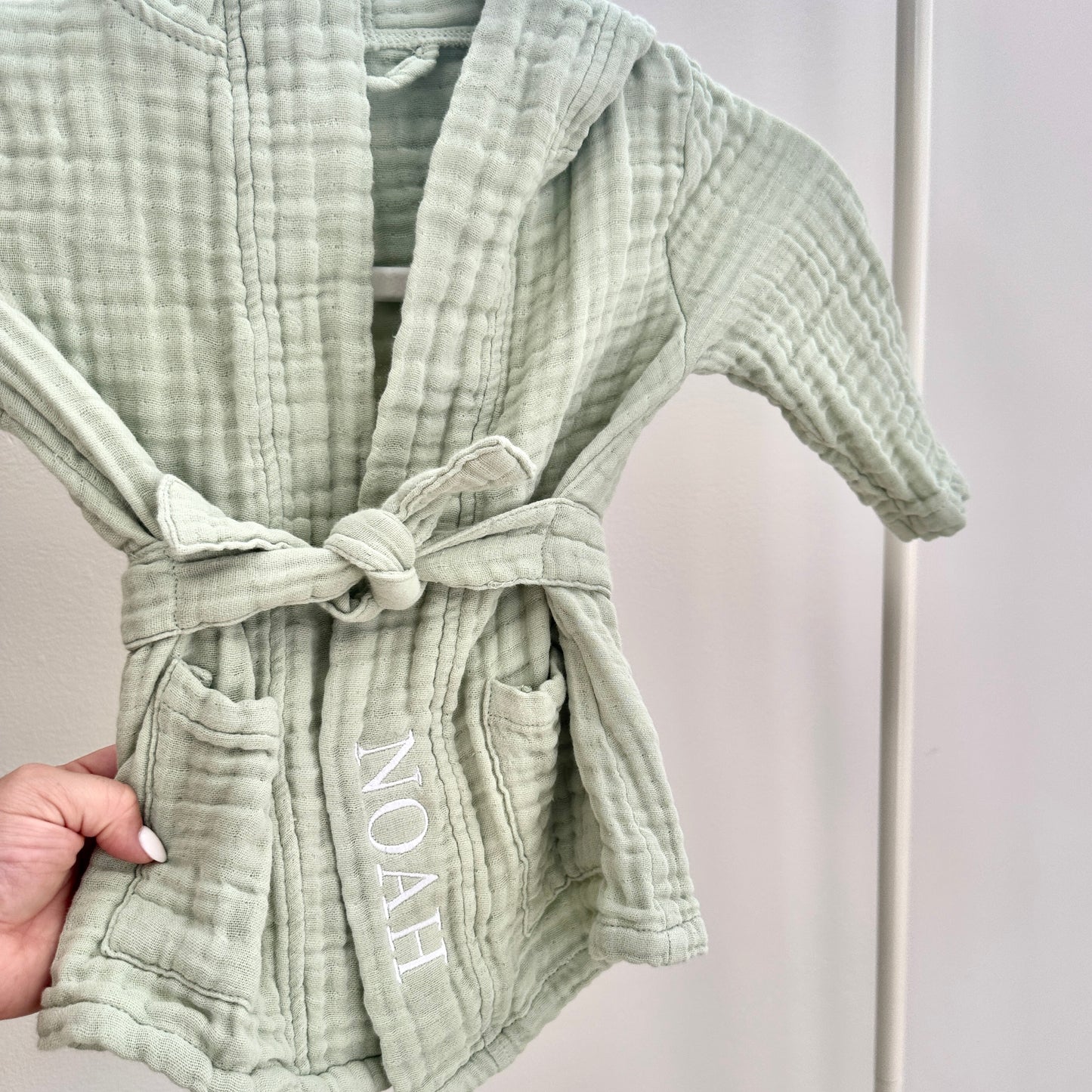 Green textured robe with 'NOAH' embroidery held by a hand against a neutral background