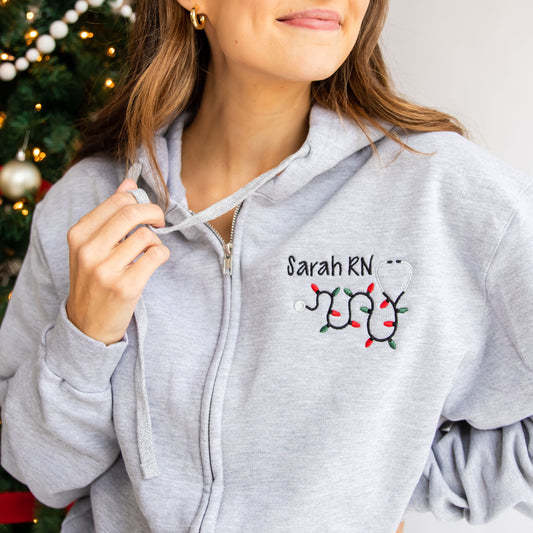 woman standing near christmas tree wearing a custom light gray full zip jacket with her name and stethoscope embroidered on to the left chest