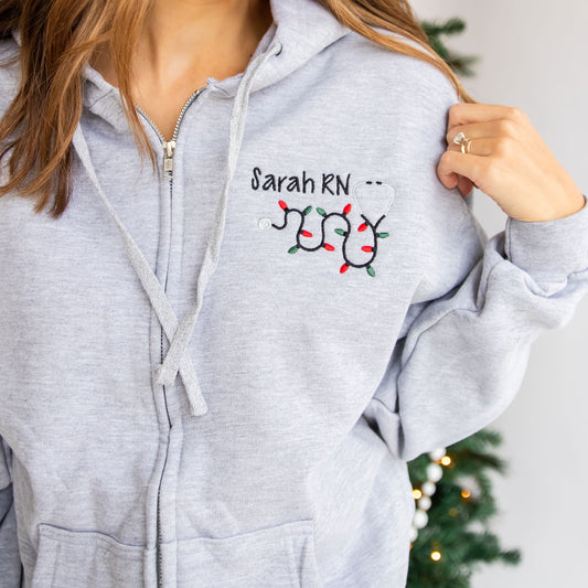 close up image of a woman's custom full zip jacket with pockets and personalized name, credentials, and christmas lights stethoscope design embroidered on the left chest