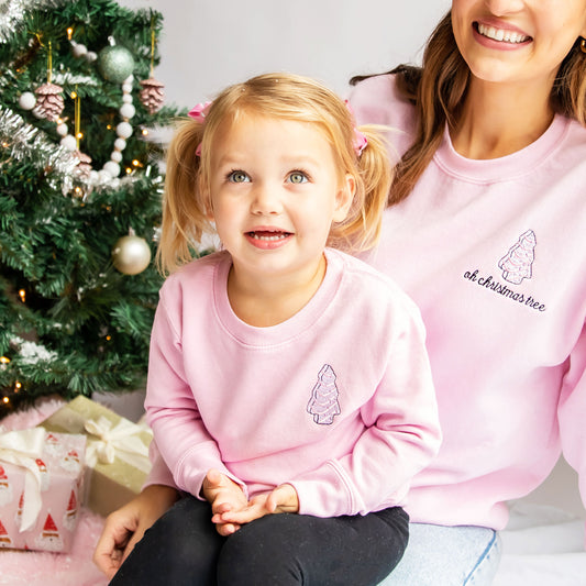 little girl sitting on her moms lap, both wearing light pink crewneck sweatshirts with little debbie christmas trees embroidered on the left chest