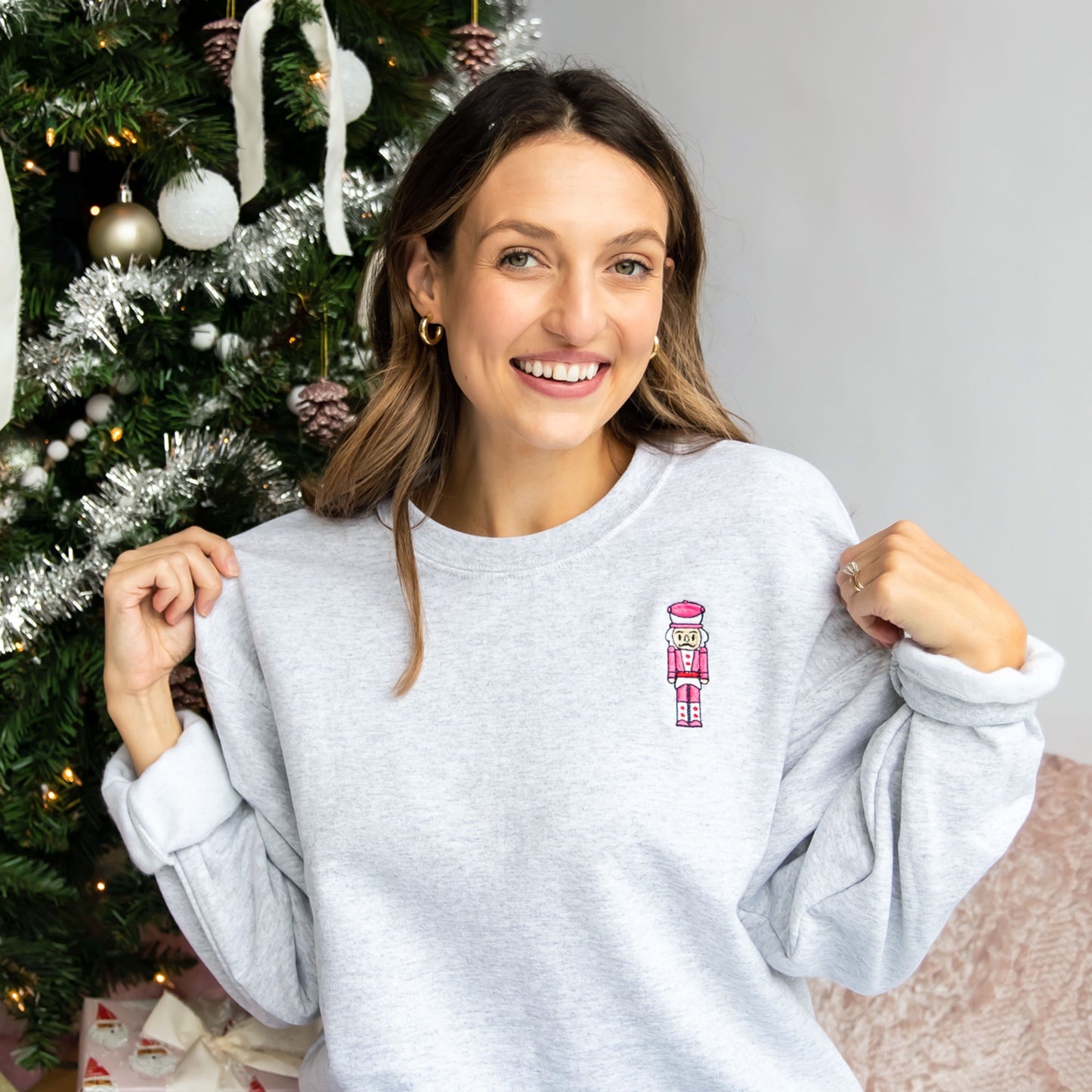 Woman styling an ash crewneck sweatshirt that features a small pink and red embroidered nutcracker on the left chest. 