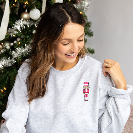 Woman wearing an Ash crewneck sweatshirt featuring an embroidered pink and red nutcracker on the left chest. 