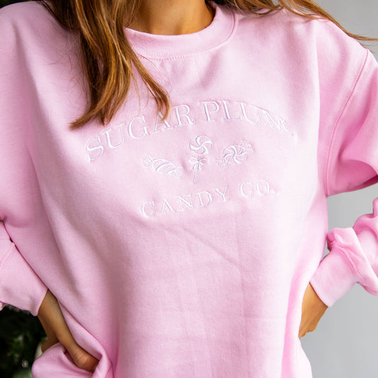 Close up of a woman wearing a pink crewneck sweatshirt that has the words Sugar Plum Candy Co embroidered across the chest in white thread.