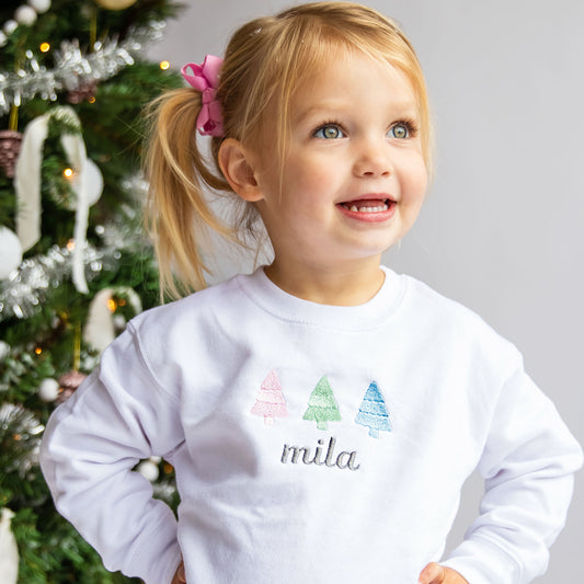 little girl wearing a white crewneck sweatshirt with three mini trees embroidered in light pink, sage, and light blue thread across the chest with her name in a dark gray thread underneath