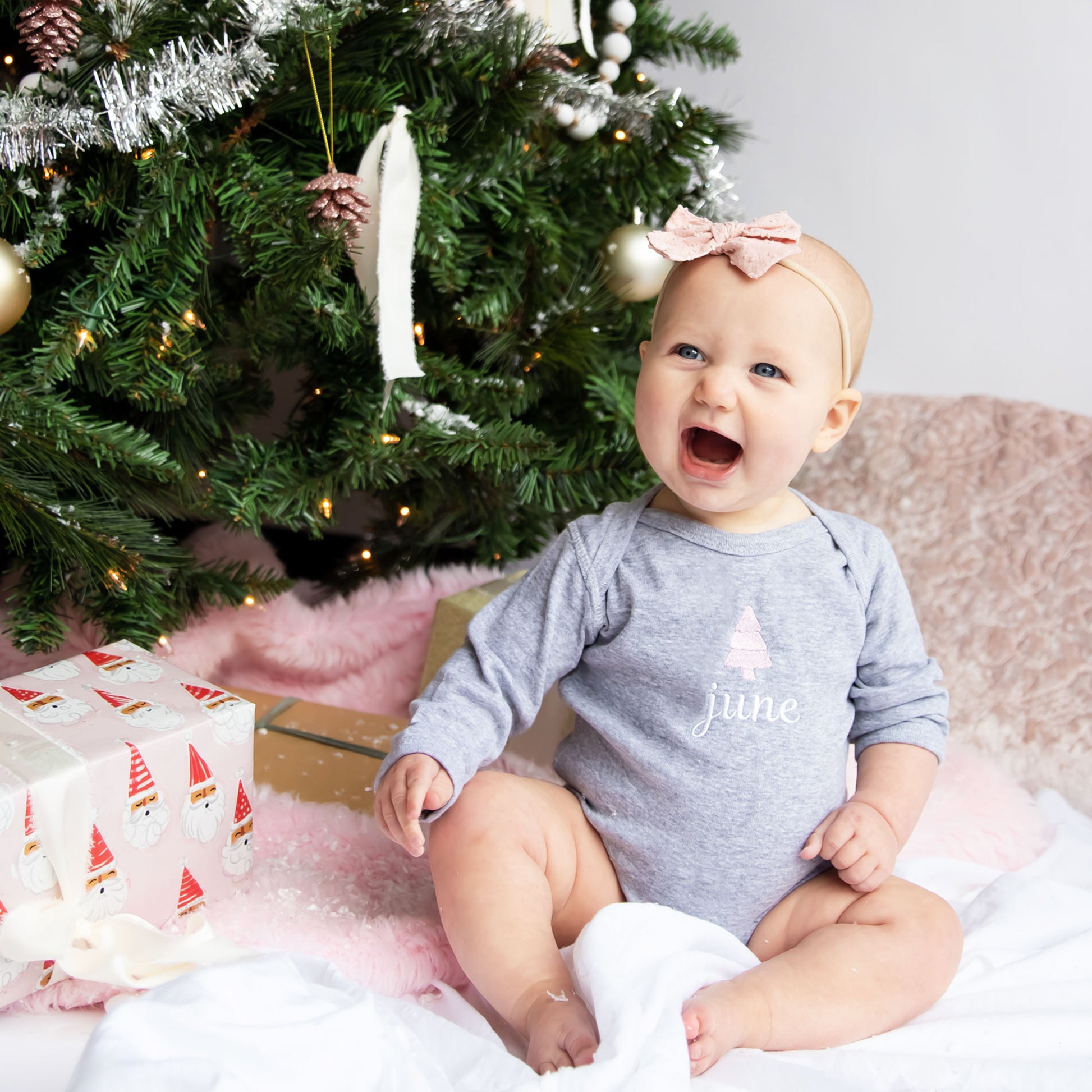 baby girl sitting near a christmas tree with presents under the tree wearing a heather gray long sleeve onesie with a custom name and mini tree embroidery design 