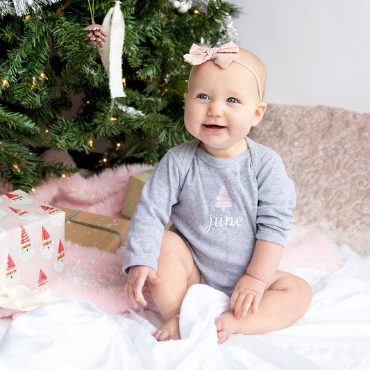 baby girl sitting by a pink decorated christmas tree wearing a gray bodysuit with personalized name and mini tree embroidery