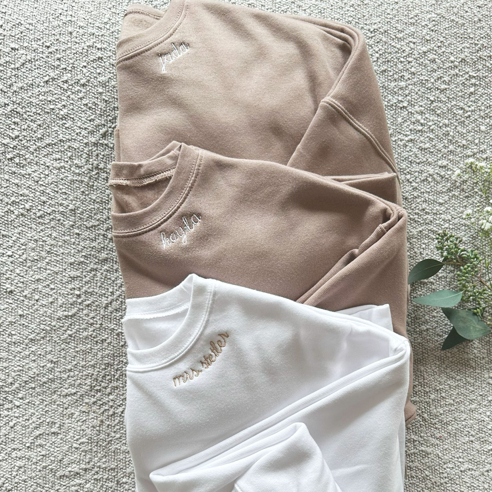 two tan crewneck sweatshirts with white neckline embroidery and a white crewneck sweatshirt with neckline name embroidered in camel
