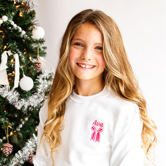 young girl standing in front of a christmas tree in a white crewneck sweatshirt with a personalized embroidered name and ribbon bow design on the left chest