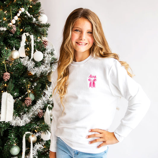 little girl wearing a white crewneck sweatshirt with a custom name and bow embroidered design on the left chest