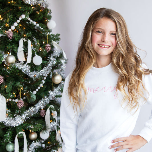 little girl wearing a white crewneck sweatshirt with merry embroidered across the chest in a script font