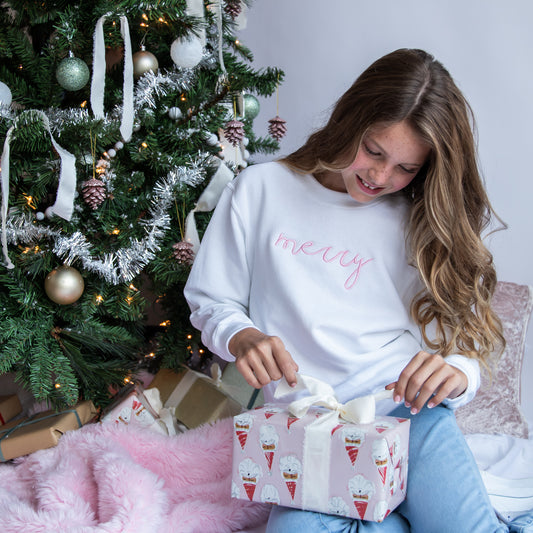little girl sitting in front of a christmas tree opening a gift and wearing a white crewneck sweatshirt with merry embroidered across the chest in a script font and baby pink thread