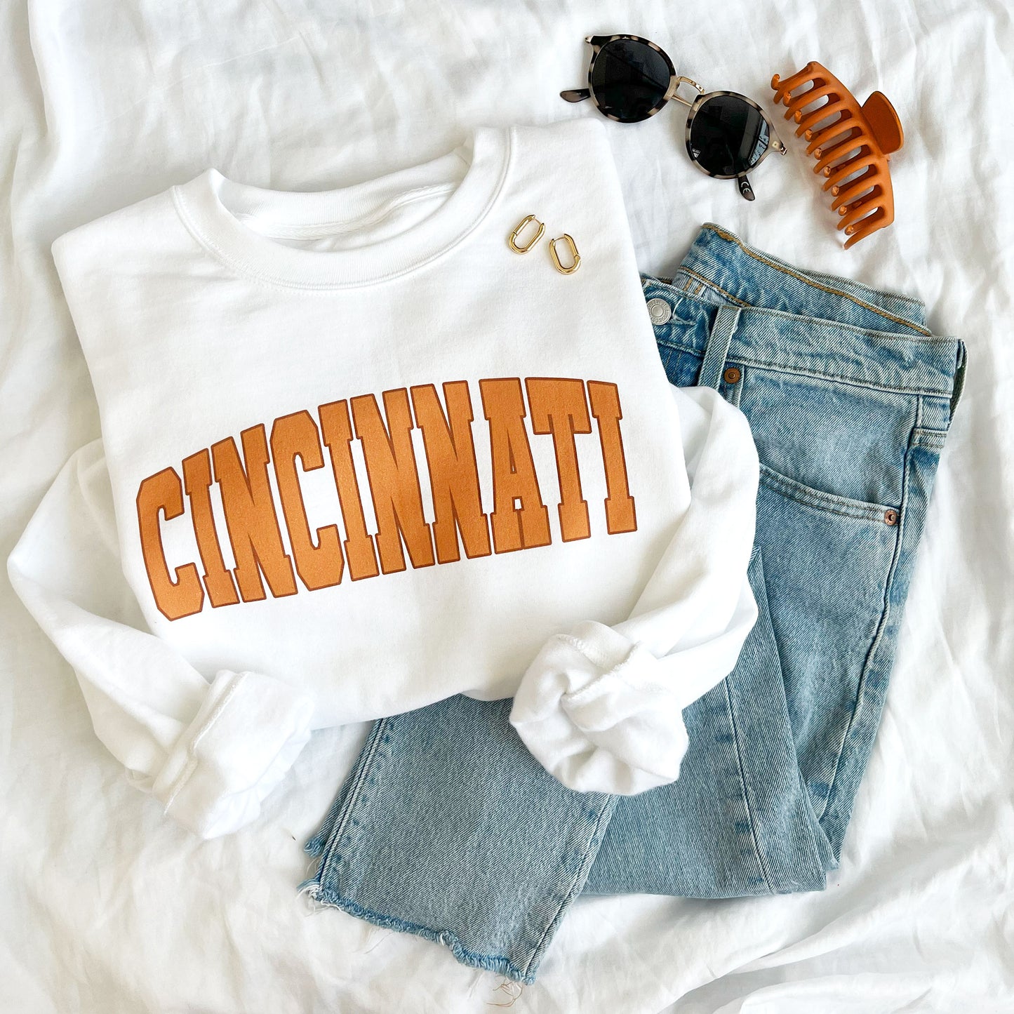 flat lay of a white crewneck sweatshirt with printed cincinnati design in orange ink styled with jeans, sunglasses, earrings, and a hair clip