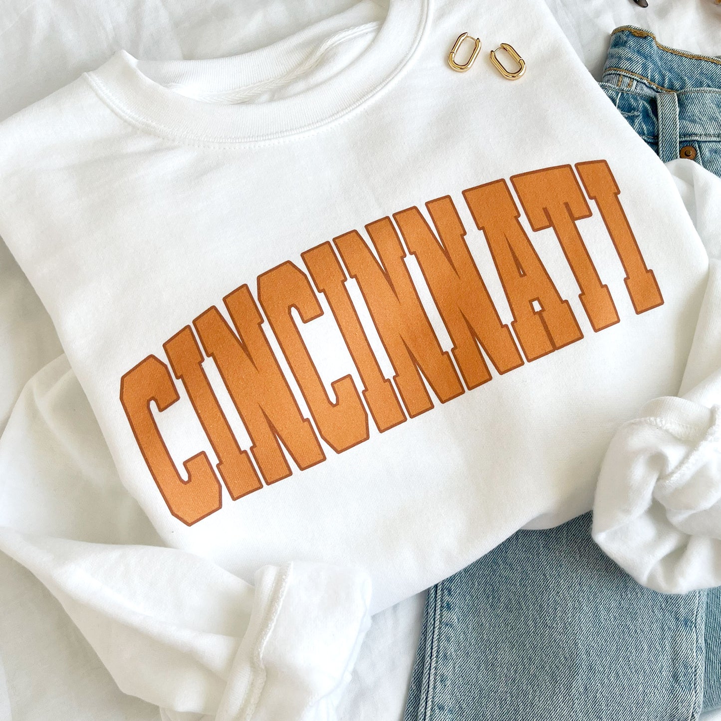 close up of a flat lay of a white crewneck sweatshirt with printed cincinnati design in orange ink 