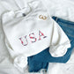 flat lay of a white crewneck sweatshirt with embroidered USA floral design in red, white and navy threads styled with jeans an gold hoop earrings