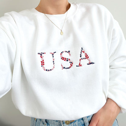 close up of a woman wearing a white crewneck sweatshirt with embroidered USA floral design in red, white and navy threads.