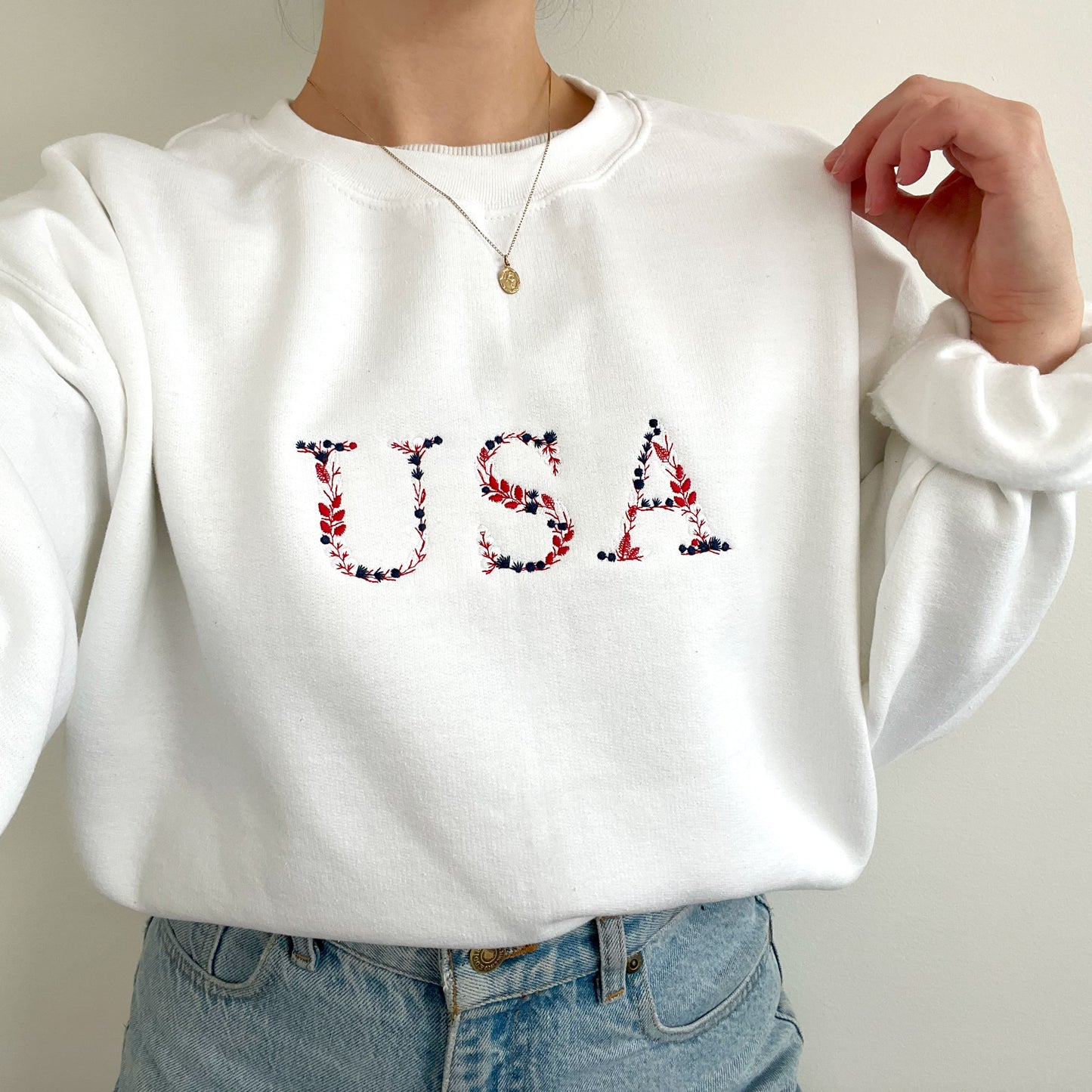 woman wearing a white crewneck sweatshirt with embroidered USA floral design in red, white and navy threads.