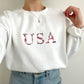 woman wearing a white crewneck sweatshirt with embroidered USA floral design in red, white and navy threads.