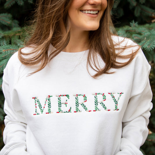 Woman wearing a white crewneck sweatshirt featuring an embroidered floral MERRY in Christmas colors. 