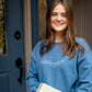 girl standing in front of a door going to a holiday party wearing a blue sweatshirt with oh holy night embroidered across the chest in a delicate script font