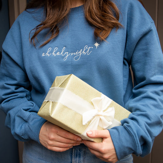 woman holding a wrapped christmas present wearing an indigo blue sweatshirt with oh holy night embroidered across the chest