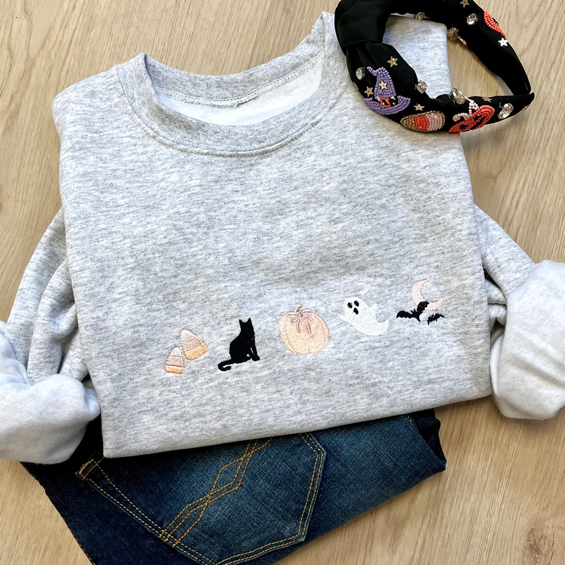 Ash sweatshirt with Halloween-themed embroidered icons ( candy corn, black cat, pumpkin with bow, ghost, moon and bats) paired with dark wash jeans and a Halloween themed headband. 