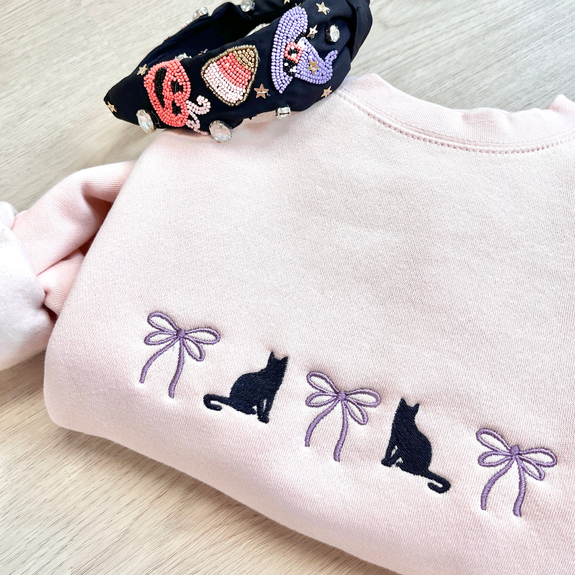 Blush pink sweatshirt with black cat and bow embroidered design across the chest, paired with a  decorative Halloween headband.