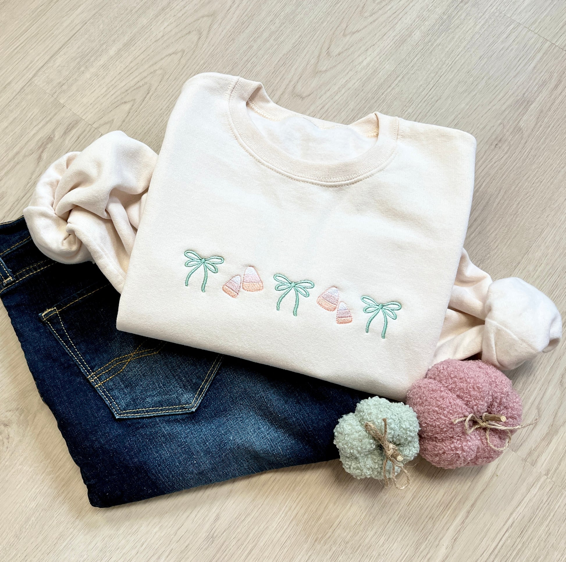 Folded sweet cream sweatshirt with embroidered bow and candy corn design across the chest laying  on a wood floor. Sweatshirt paired with jeans and decorative pumpkins.