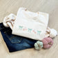 Folded sweet cream sweatshirt with embroidered bow and candy corn design across the chest laying  on a wood floor. Sweatshirt paired with jeans and decorative pumpkins.