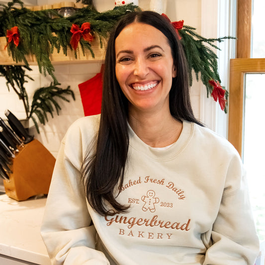 woman in her kitchen wearing a sand crewneck sweatshirt with a gingerbread bakery embroidered design across the chest