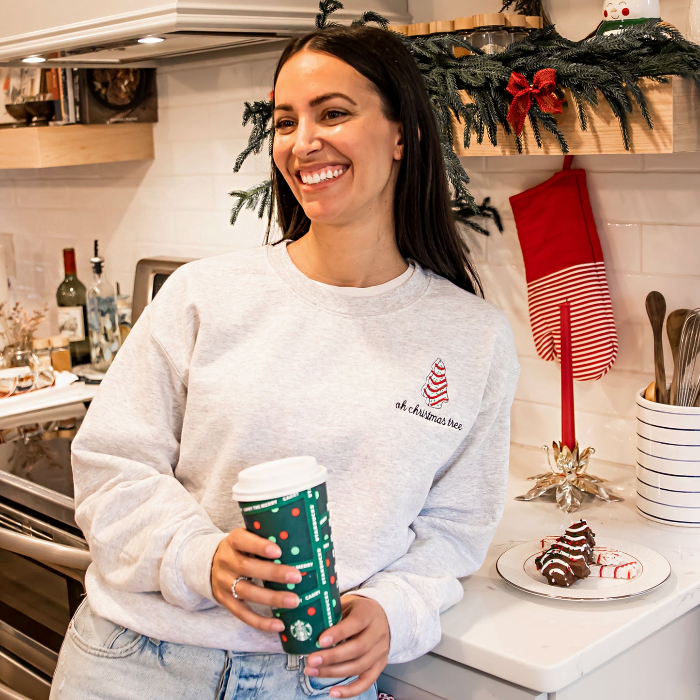 Woman styling an ash crewneck sweatshirt in a Christmas decorated kitchen. The sweatshirt features an embroidered little debbie Christmas tree and the text oh christmas tree below.
