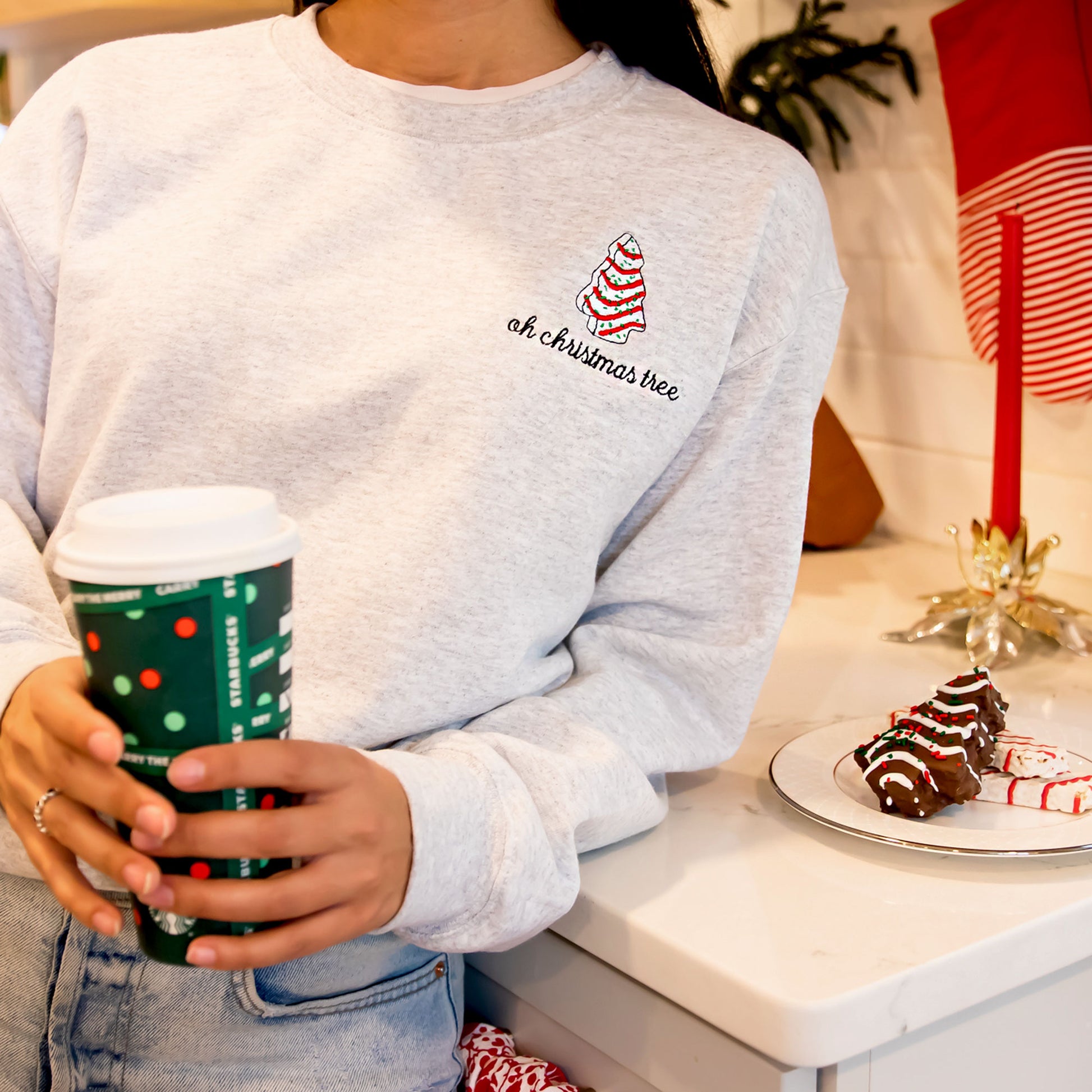 Woman wearing an ash crewneck sweatshirt holding a Starbucks cup and leaning on a kitchen counter. The sweatshirt has the embroidered little Debbie Christmas tree and the text oh Christmas tree underneath.