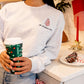 Woman wearing an ash crewneck sweatshirt holding a Starbucks cup and leaning on a kitchen counter. The sweatshirt has the embroidered little Debbie Christmas tree and the text oh Christmas tree underneath.
