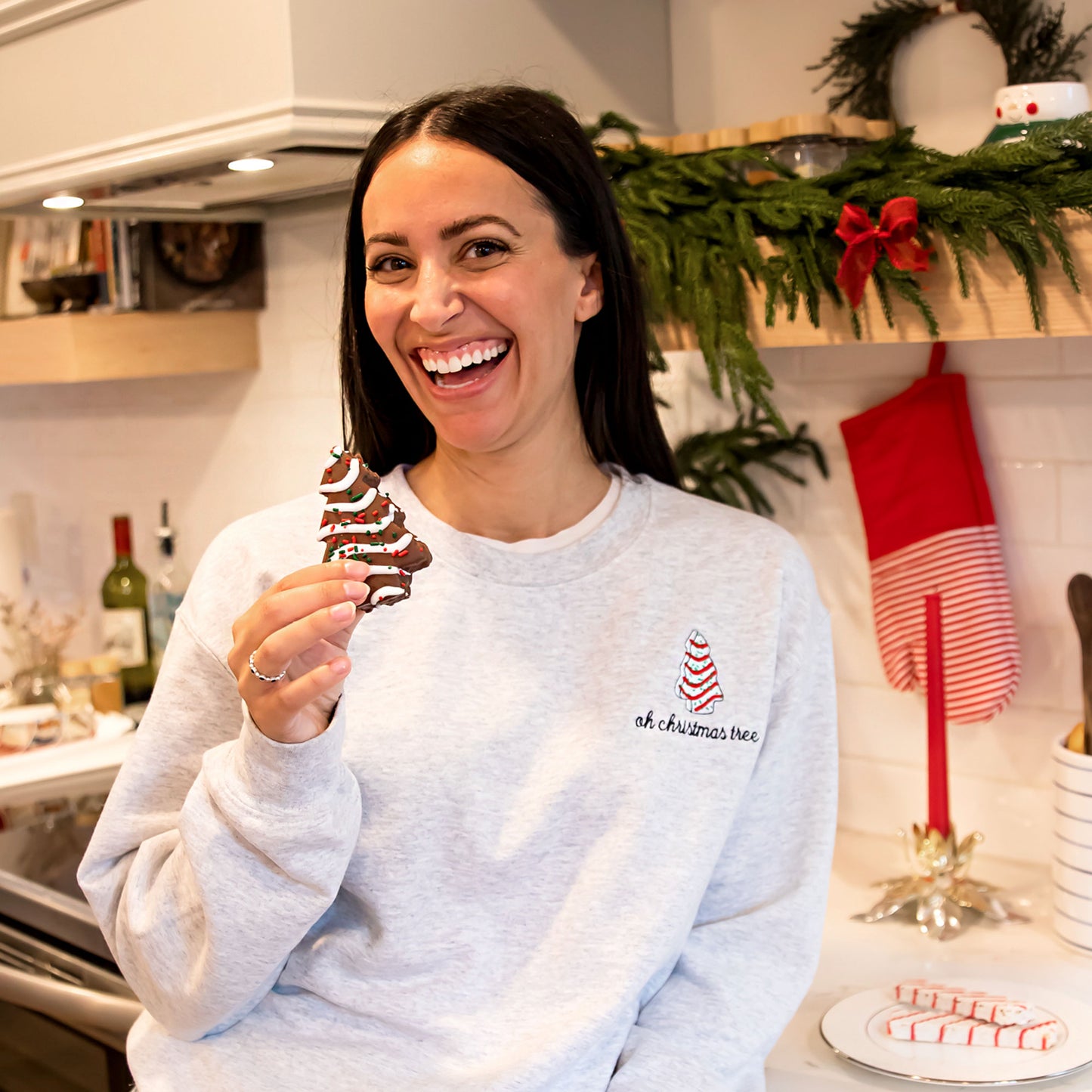 Woman holding a little Debbie Christmas tree and modeling an ash crewneck sweatshirt with a little debbie embroidered design and the words  Oh Christmas tree underneath the design. 