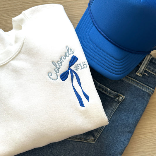 Folded white shirt with 'Colonels #15' and long tail bow embroidery on the left chest, blue cap, and jeans 