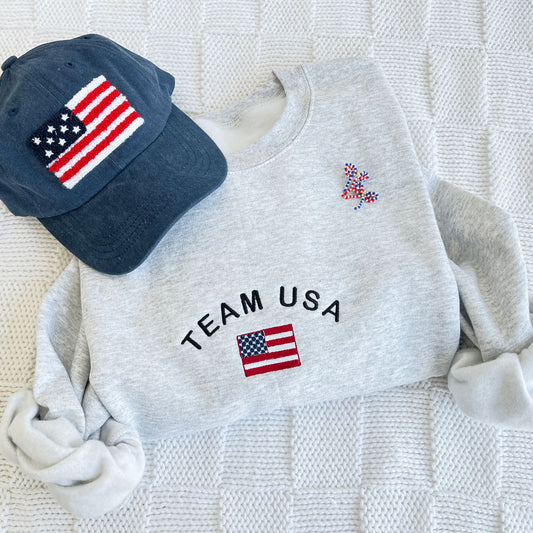 ash crewneck sweatshirt with team usa and a mini american flag embroidered on the center chest paired with a navy baseball cap with a chenille usa patch and red white and blue bow earrings