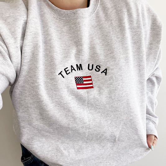 woman wearing an ash crewneck sweatshirt with team usa and mini american flag embroidered on the center chest