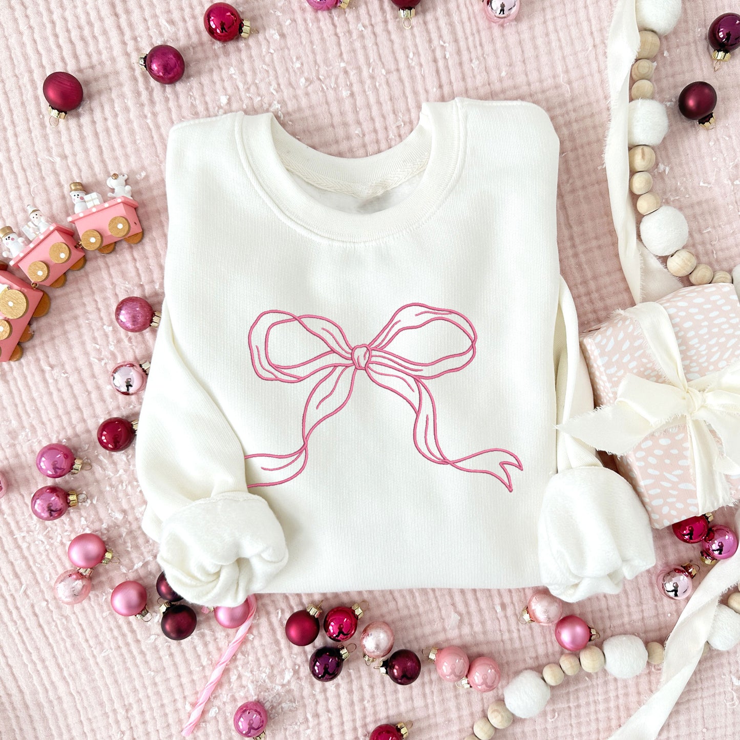 Christmas styled flat lay of a sweet cream crewneck with embroidered ribbon bow design in pink thread large across the chest