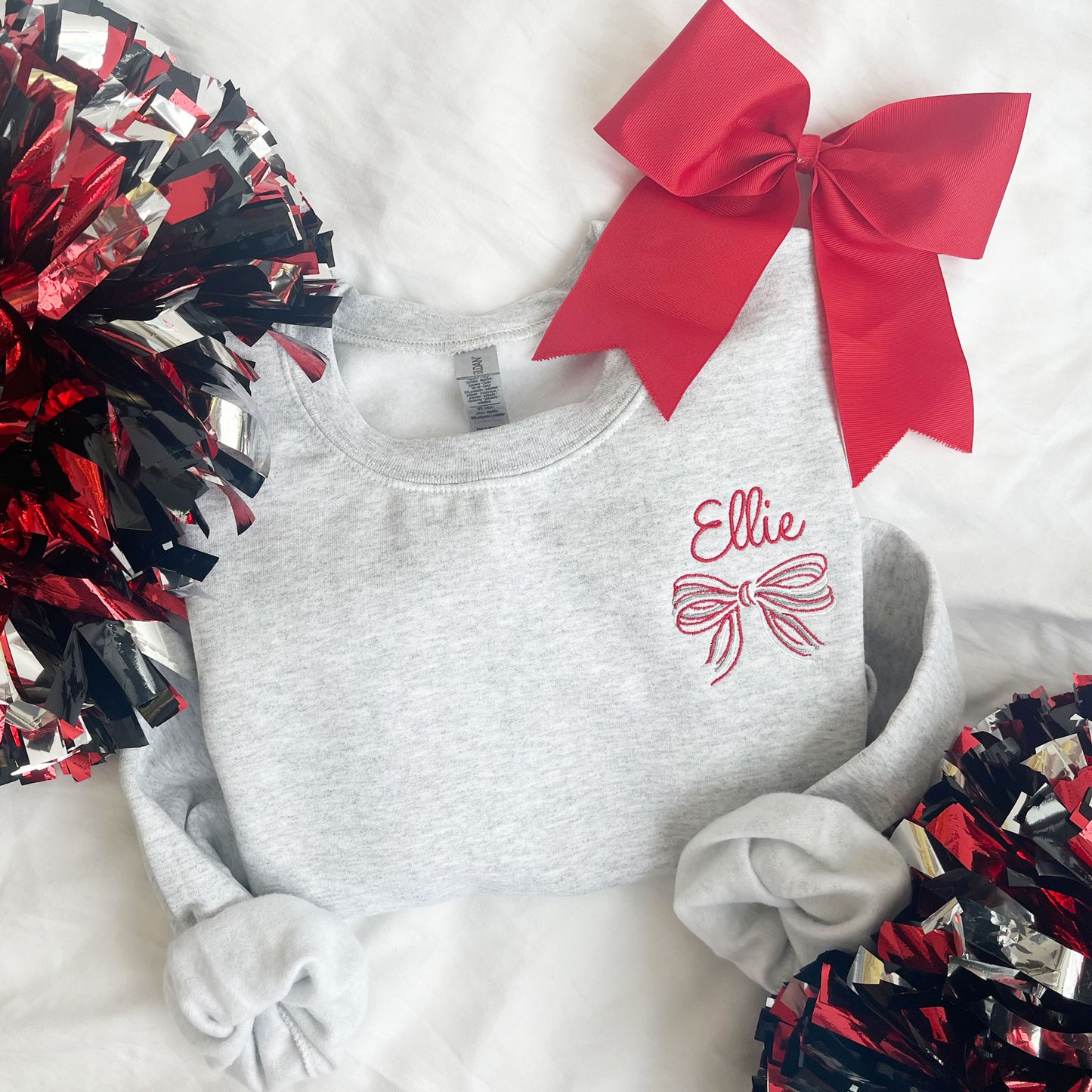 ash crewneck sweatshirt with a custom name and striped bow embroidered design on the left chest surrounded by pom poms and large bow