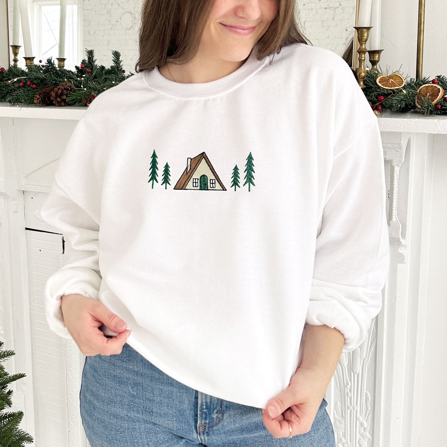 young woman wearing a white crewneck sweatshirt with embroidered a frame cabin design across the chest