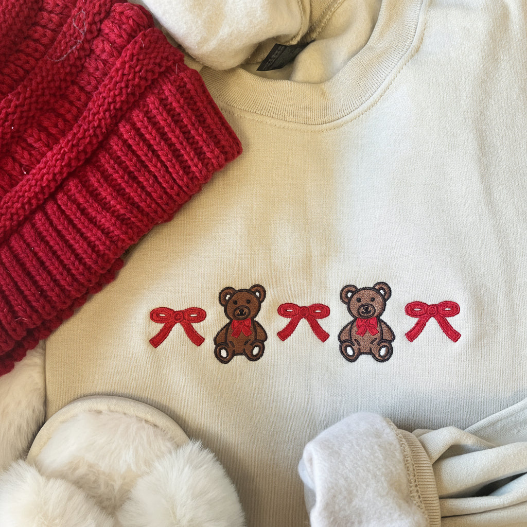 Beige sweatshirt with embroidered teddy bears and red bows, surrounded by red knitted item and white fabric.