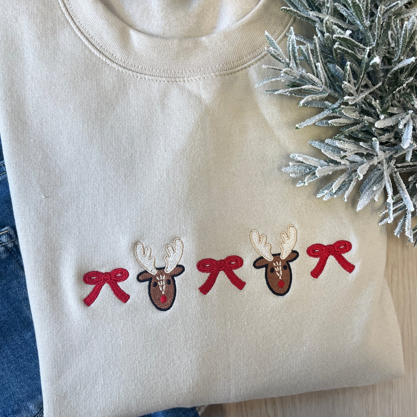 Beige sweatshirt with reindeer and bow designs, next to a sprig of frosted branches.