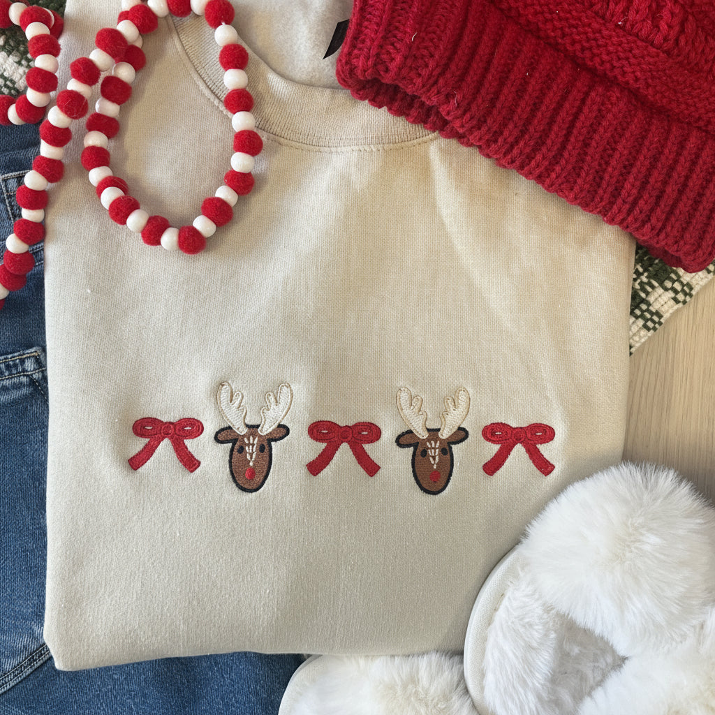 Sand crewneck sweatshirt with reindeer and bow embroidered design, surrounded by festive items like a red knitted hat and white fuzzy slippers.