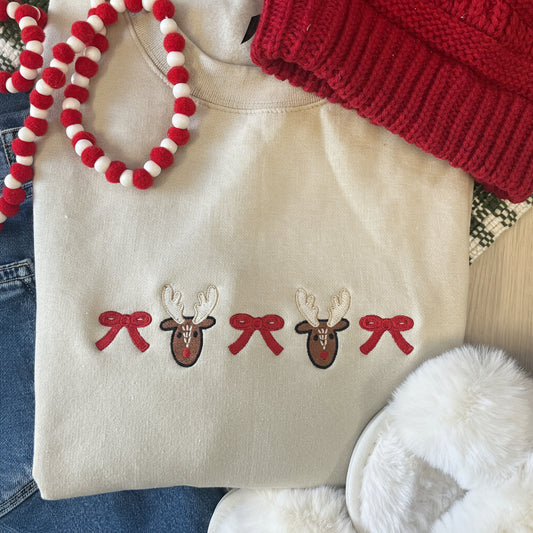 Sand crewneck sweatshirt with reindeer and bow embroidered design, surrounded by festive items like a red knitted hat and white fuzzy slippers.