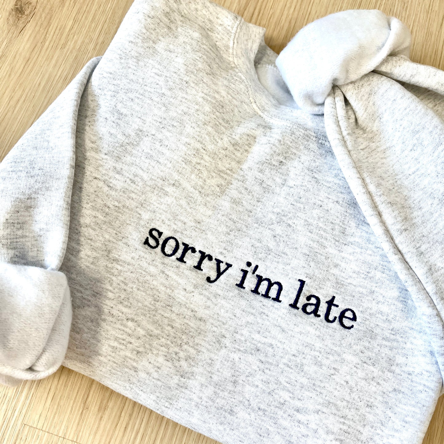 Gray sweatshirt with 'sorry i'm late' embroidered in navy thread on a wooden surface