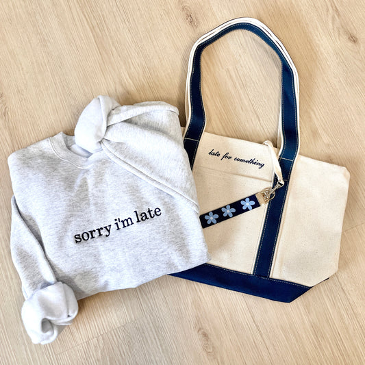 Gray sweatshirt with 'sorry i'm late' text and white tote bag with blue accents on a wooden surface