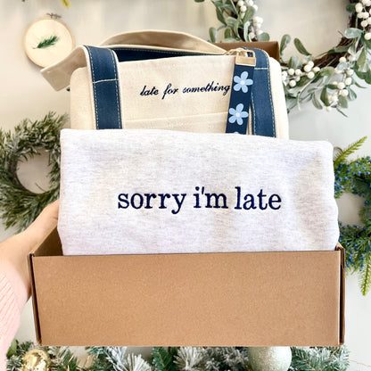 Box containing a towel with 'sorry i'm late' text and a tote bag with 'late for something' text, surrounded by festive decorations.